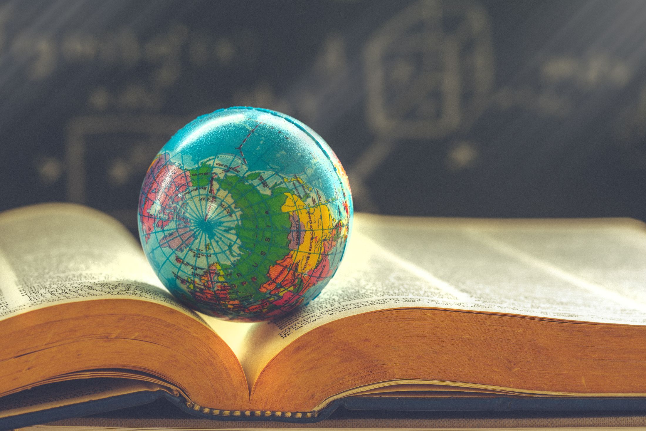 Small colorful globe resting on an open book against a blurred chalkboard background.