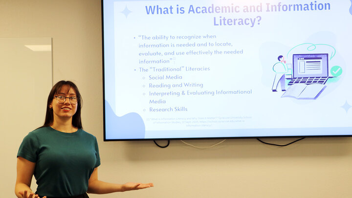 A student stands next to a white presentation screen as she presents on academic literacy in a class.